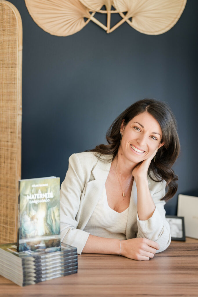 Céline Salvado assise à son bureau, souriante, à côté d’une pile de livres *La Maternité Consciente*, incarnant la sérénité et la passion qui animent son travail d’autrice.